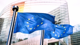 Zwei EU-Flaggen im Wind wehend, im Hintergrund Gebäude mit Glasfassade