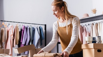 Person hält gefaltete Kleidung und bereitet diese für Versand vor, im Hintergrund befinden sich Kleiderstangen mit weiteren farbenfrohen Kleidungsstücken
