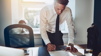 Person in Businesskleidung steht vor einem Schreibtisch und unterzeichnet ein Dokument, daneben steht eine Aktentasche sowie ein Glas Wasser 