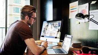 Person mit Brille und kurzen braunen Haaren sitzt an einem Schreibtisch mit Monitor und Laptop und blickt auf Bilder am Bildschirm während Bücher sowie Kopfhörer am Tisch liegen und auf der Wand Infoblätter befestigt sind
