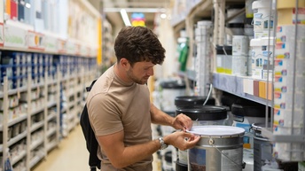 Seitliche Aufnahme einer Person, die in einem Gang vor einem Eimer steht und den Deckel des Eimers leicht anhebt. Links und rechts im Gang sind lange, hohe Regale, die mit weiteren Eimern befüllt sind.