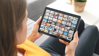 Blick über die Schulter einer Person mit gelben Pullover auf ein Tablet mit Bilder-Kacheln am Bildschirm, das die Person mit beiden Händen hält