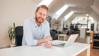 Lächelnde Person mit rötlichen kurzen Haaren, Bart und blauem Hemd sitzt an einem Schreibtisch mit Laptop, Notizbuch und Stift in einem großen Büro