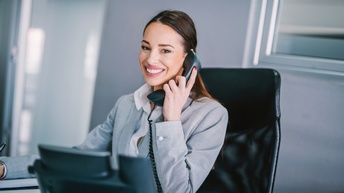 Lächelnde Person mit dunklen geschlossenen Haaren und Businesskleidung telefoniert während sie bei einem Schreibtisch in einem Büro sitzt
