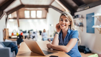 Lächelnde Person mit langen blonden Haaren und kurzer blauen Bluse sitzt an einem Schreibtisch mit Laptop in einem offenen Büroraum