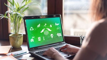 Person sitzt vor aufgeklapptem Notebook, auf dessen Monitor auf grünem Hintergrund weiße Symbole zum Thema Nachhaltigkeit aufscheinen