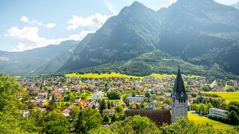 Ortschaft in einem Tal vor hohen, bewaldeten Bergen. Im Vordergrund stehen Bäume und eine Kirche mit einem hohen, spitzen Turm. Dahinter erstrecken sich zahlreiche Wohnhäuser und weitere Gebäude über die Talfläche