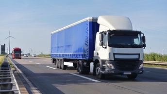 Weiß-blauer LKW fährt auf einer Autobahn, im Hintergrund zeigen sich Windräder