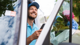 Lächelnde Person mit Kappe, lugt vom Autofahrersitz zwischen Auto und geöffneter Türe hervor, hält in einer Hand Smartphone und in anderer Blumenstrauß mit pinken und blauen Hortensien