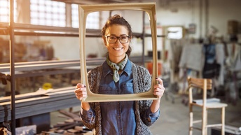 Lächelnde Person in Werkstatt stehend hält leeren Rahmen in Händen und lugt hindurch