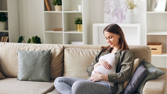 Eine Person mit gekreuzten Beinen sitzt auf einem beigen Sofa. Ihre Hände liegen auf ihrem nach außen gewölbten Bauch, auf den sie auch blickt.  Hinter ihr sind Regale mit Pflanzen, Büchern und Bilderrahmen