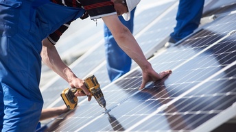 Person in blauer Arbeitslatzhose mit weißem Helm mit Bohrmaschine in der Hand beim der Montage von Solarpanelen