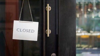 Ein Schild mit dem Wortlaut closed hängt an einer geschlossenen dunklen Tür aus Holz und Glas. Im rechten Bildausschnitt sieht man verschwommen durch eine Glasscheibe das Innere des Gebäudes bestehend aus Tischen, Sesseln und Lampen