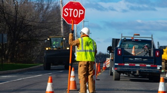 Eine Person mit einer Warnweste und Helm steht auf einer Straße und hält ein Stoppschild, im Hintergrund abgestellte Autos und ein fahrender Bagger.