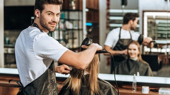 Person in Schürze föhnt Haare weiterer Person vor ihr mit Haarschneideumhang sitzend, im Hintergrund verschwommen großer Spiegel