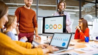 Personen an einem Tisch in einem modernen Büro unterhalten sich während Analysen und Grafiken auf einem Bildschirm sowie auf einem Laptop visualisiert sind