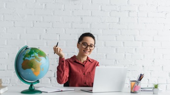 Person mit geschlossenen dunklen Haaren und Brille sowie roter Bluse sitzt bei einem Schreibtisch und blickt freudig darauf während ein Stift in der Hand gehalten wird, am Tisch steht ein Globus, ein Stiftebecher sowie weitere Unterlagen liegen darauf