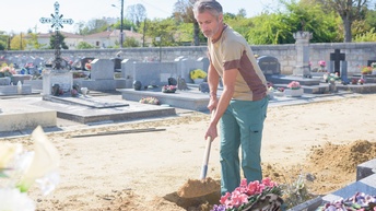 Eine Person gräbt mit einer Schaufel Erde um, sie hebt eine Grube für ein Grab in einem Friedhof aus. Ringsherum sind bunte Blumengestecke und Gräber, man sieht eine Mauer, Häuser und Bäume im Hintergrund des Friedhofs. 