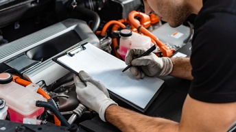 Seitliche Aufnahme einer Person, die sich mit den Unterarmen am Rand einer geöffneten Motorhaube abstützt. Die Person trägt Handschuhe und notiert etwas auf einem Klemmbrett