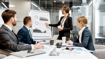 Personen in einem Besprechungsraum blicken gespannt auf eine analytische Chart, während auf ihrem Schreibtisch Laptops, Kaffeebecher sowie Statistiken liegen