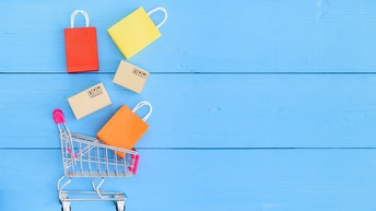 Auf einer blauen Unterfläche liegt in der linken Hälfte ein Miniatur-Einkaufswagen. Über dem Einkaufswagen liegen drei bunte Einkaufstaschen und zwei Kartons. Auf einem Karton steht fragile.