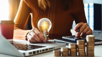 Auf einem Tisch sind fünf, unterschiedlich hohe Münzstapel, ein aufgeklappter Laptop, ein Notizbuch, auf dem ein Taschenrechner ist sowie die Hände einer Person. Die rechte Hand hält eine leuchtende Glühbirne, die linke einen Stift