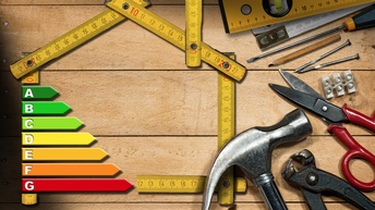 Topshot eines Maßbandes, das so drapiert ist, dass es die Umrisse eines Hauses ergibt. Links im Umriss ist ein Balkendiagramm zum Energiebedarf. Im unteren Bereich liegen verschiedene Werkzeuge wie Hammer, Bohrer, Nägel oder Zangen.