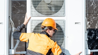 Rückenansicht einer Person mit schwarz-oranger Jacke, orangem Helm und Sonnenbrille, die neben einem Außengerät einer Klimaanlage steht und beide Hände auf dem Gerät hat