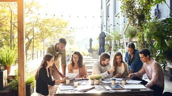 In einem überdachten Außenbereich sitzen um einen Tisch fünf Personen. Daneben stehen zwei weitere Personen. Auf dem Tisch liegen verschiedene Unterlagen. Die Personen blicken auf diese und unterhalten sich in zwei Gruppen