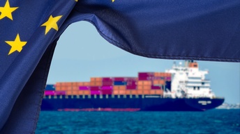 Im Vordergrund ist die Flagge der Europäischen Union, wie ein Vorhand zur Seite geschoben. Dahinter ist ein Containerschiff beladen mit zahlreichen Containern auf dem Meer, um Güter zu transportieren