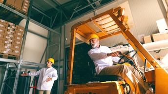 Seitliche Aufnahme einer Person mit Helm, die in einem Gabelstapler sitzt und nach hinten blickt. Hinter dem Gabelstapler steht eine weitere Person mit Helm, die die Arme vor sich ausgestreckt hat 