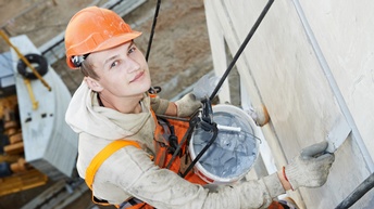 Person mit oranger Schutzkleidung sowie Schutzhelm blickt in die Kamera, hängt mit Sicherung an Hausfassade und bearbeitet diese