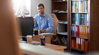 Lächelnder Unternehmer sitzt schreibend an Schreibtisch mit Bücherregal hinter sich