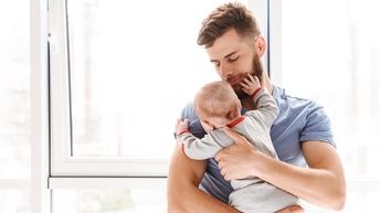 Frontalaufnahme einer Person mit kurzen Haaren und Bart, die vor einem Fenster steht. Mit beiden Händen hält die Person ein Baby vor den Oberkörper und blickt darauf