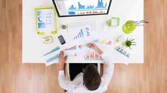 Person in Vogelperspektive an Schreibtisch sitzend auf dem Computer mit Statistik am Monitor steht, analysiert unterschiedliche Dokumente mit statistischen Diagrammen