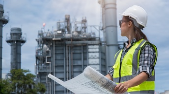 Person mit Warnweste und Schutzhelm hält mit beiden Händen einen Bauplan und blickt zu einem Fabriksgelände