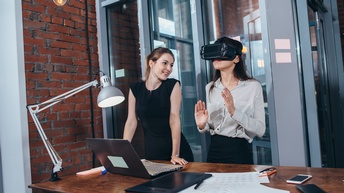 Eine Person mit Virtual Reality Brille steht an einem Schreibtisch während eine weitere Person neben ihr steht und diese beobachtet