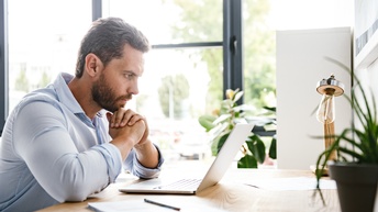 Person mit blauem Hemd und Bart sitzt konzentriert an einem Schreibtisch und blickt konzentriert auf einen Laptop während im Hintergrund Pflanzen in einem hell erleuchteten Raum mit Glasfensterfront stehen