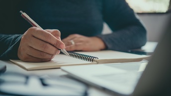 Eine Person sitzt an einem Tisch und hält einen Stift in der rechten Hand. Vor der Person liegt ein Notizbuch. Daneben ist ein aufgeklappter Laptop