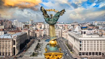 Rückenansicht einer hohen, vergoldeten Säule auf einem großen Platz. Um den Platz sind verschiedene Gebäude. Auf den Straßen fahren Autos. Im Hintergrund ist ein Regenbogen