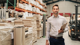 Porträt einer Person mit Klemmbrett in der rechten Bildhälfte. Links neben der Person sind viele, eher schmale übereinander gestapelt. Im Hintergrund ist in der linken Hälfte ein großes, hohes Regal,  in dem verschiedene Objekte sind und ein Gabelstapler.
