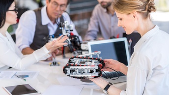 Personen sitzen an einem Tisch und betrachten Robotermaschinen, daneben stehen ein Laptop, ein Tablet sowie weitere Unterlagen