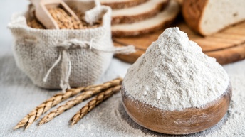 Holzschüssel mit gehäuftem Mehl, nebenliegend goldgelbe Weizenähren, im Hintergrund verschwommen Jutesack voller Weizen mit Holzschaufel sowie Holzbrett mit geschnittenem Brot