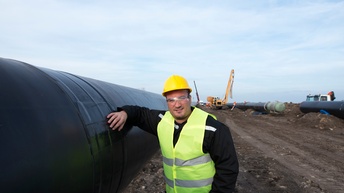 Person mit Schutzgläsern, gelbem Schutzhelm und gelber Schutzweste lehnt an großem Gasrohr, im Hintergrund weitere Gasrohre und Baumaschinen