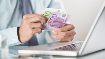 Zwei Hände einer Personen halten über einen aufgeklappten Laptop drei Geldscheine, auf dem ersten steht 500 und er ist lila, der zweite ist grün. Der dritte ist orange.