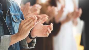 Nahaufnahme der Hände einer Person, die gerade klatscht. Im Hintergrund sind weitere Personen in Unschärfe fotografiert, die klatschen