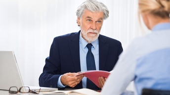 Eine Person hält die Bewerbungsmappe einer zum Bewerbungsgespräch eingeladenen Person, die gegenüber von ihr sitzt.