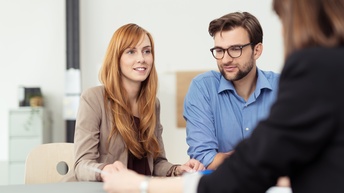 Zwei Personen sitzen an Tisch, eine Person lächelt sanft, die andere trägt Brillen, im Vordergrund verschwommen weitere Person