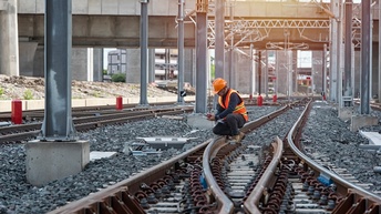 Eine Person mit Helm und Sicherheitsweste hockt auf Gleisen. Daneben sind weitere Gleise. Die Person hält ein Walkie Talkie