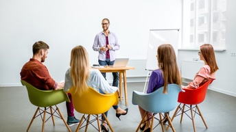 Rückenansicht von vier Personen, die in Sesseln nebeneinander sitzen. Den Personen gegenüber steht eine weitere Person hinter einem Tisch. Neben der Person ist ein Whiteboard.
